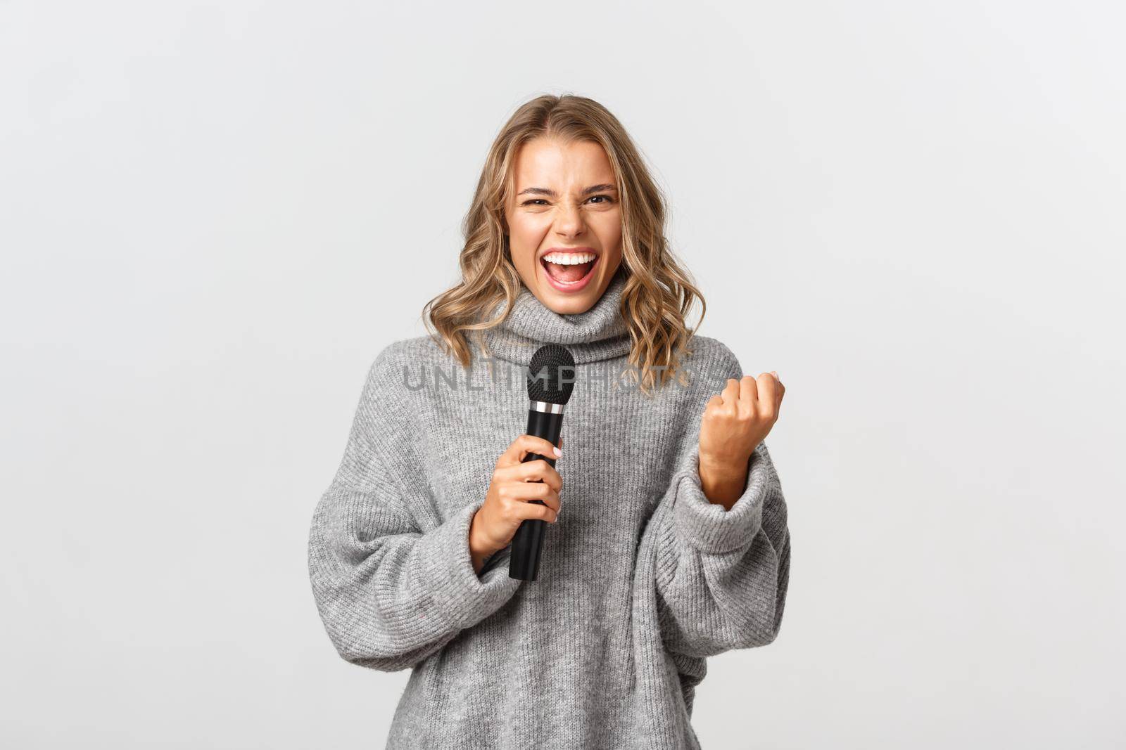 Sassy beautiful girl in grey sweater, looking excited and singing in microphone, standing over white background.