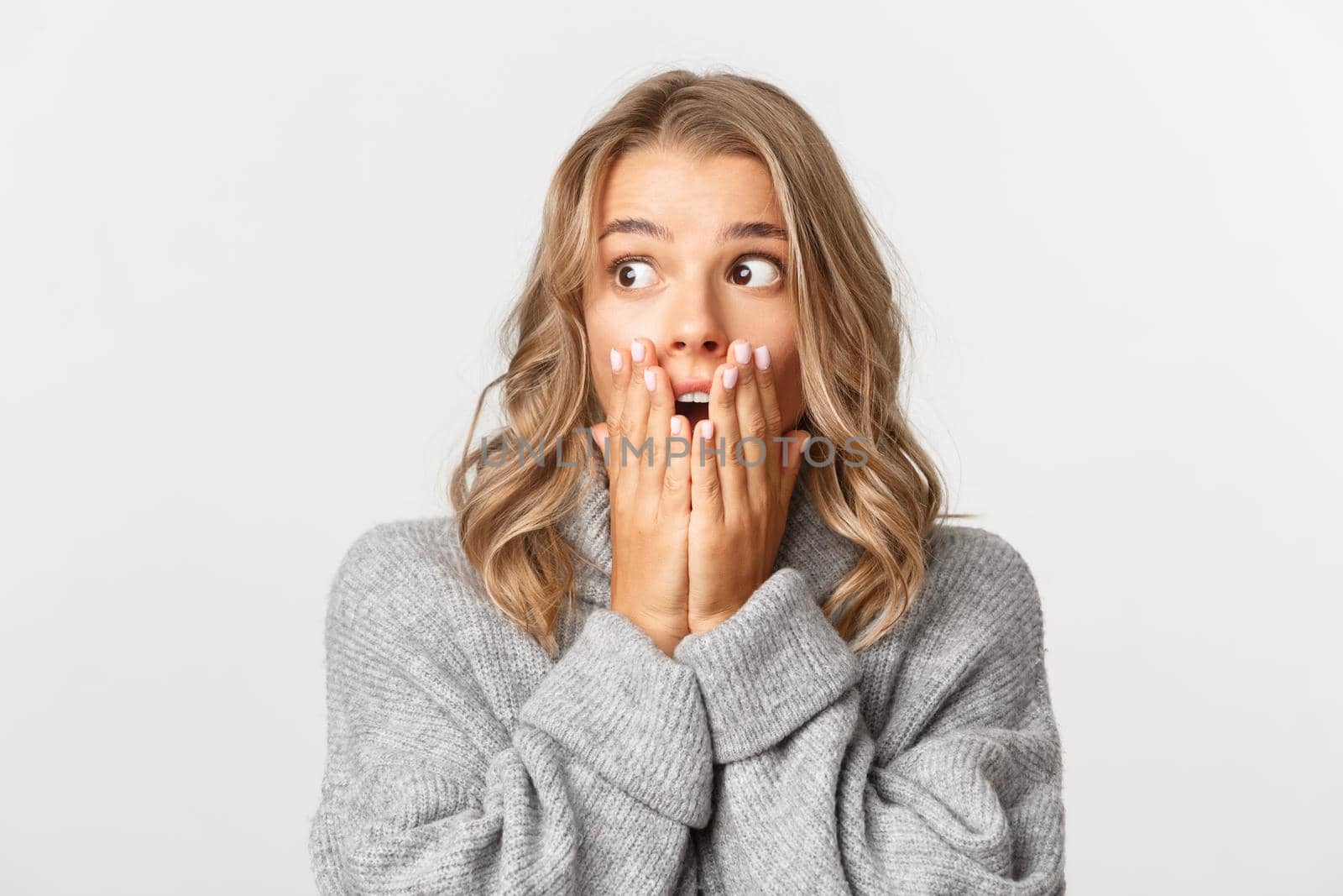 Close-up of shocked and surprised blond girl in grey sweater, looking at upper left corner, gasping amazed, standing over white background.