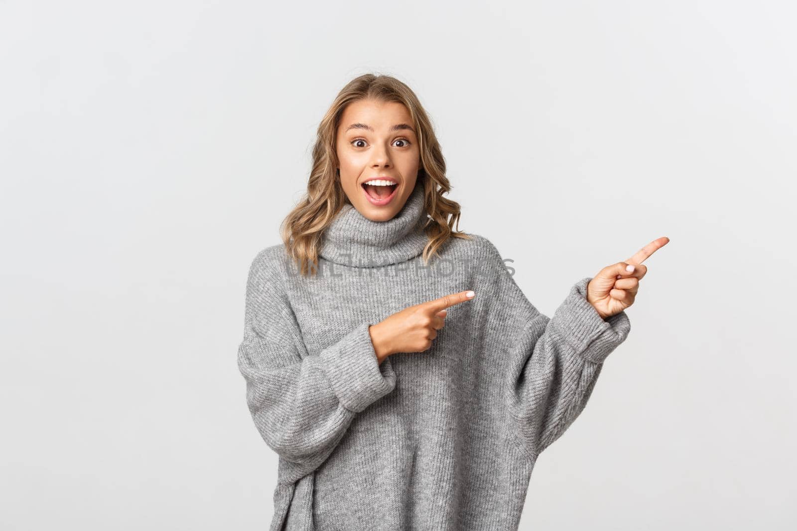 Image of amazed blond girl showing your logo, pointing fingers right and smiling excited, standing over white background.