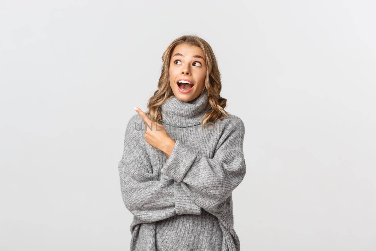 Portrait of amazed blond girl in grey sweater, open mouth and looking fascinated at your logo, pointing finger at upper left corner, white background.