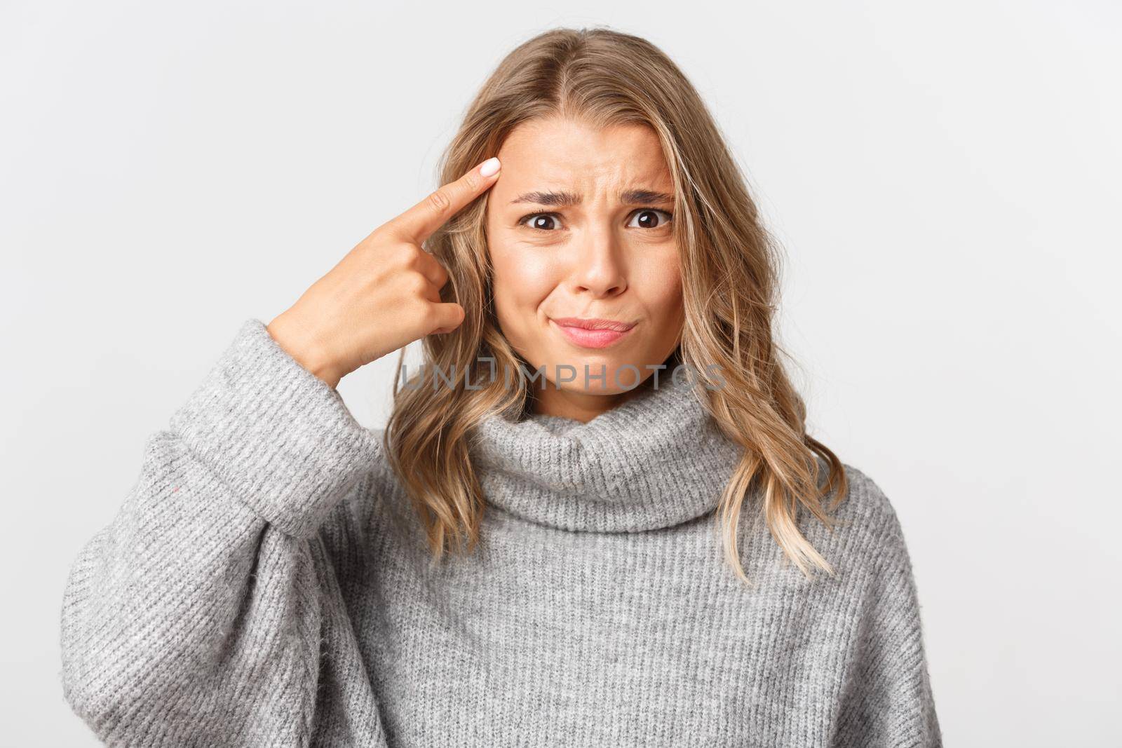 Close-up of confused blond girl in grey sweater, pointing finger at head, scolding something for acting stupid, standing over white background.