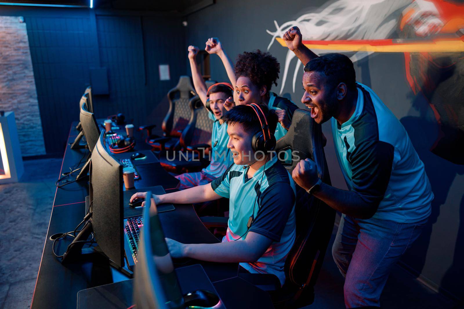 Multiracial team of happy professional cyber sports gamers celebrating success wile raising hands up during esports tournament in gaming club