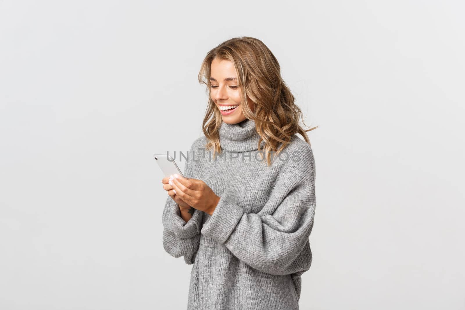 Image of attractive caucasian girl in grey sweater laughing at message, looking at mobile phone and smiling, standing over white background.