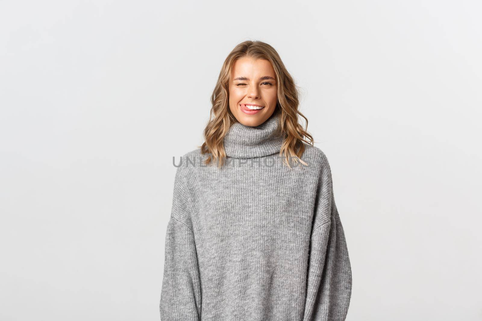 Sassy attractive blond girl in grey sweater winking, showing tongue and flirting with someone, standing over white background.