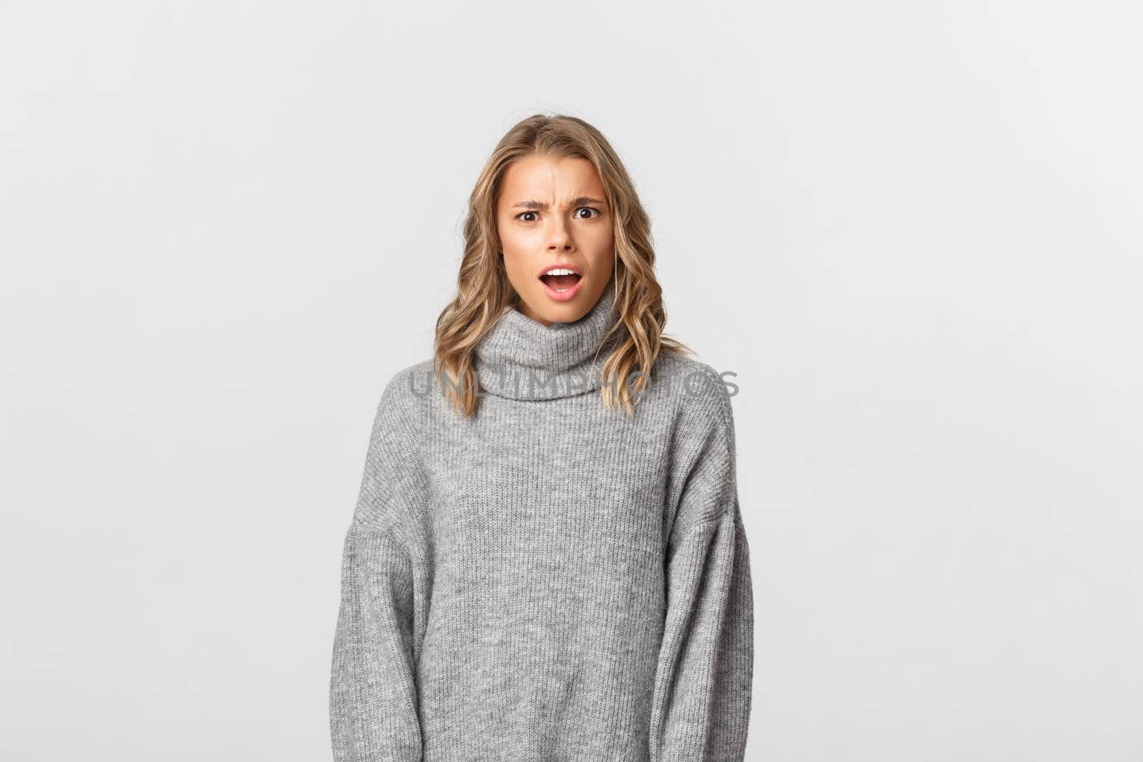Close-up of confused blond girl in grey sweater, open mouth and looking puzzled at camera, standing over white background by Benzoix