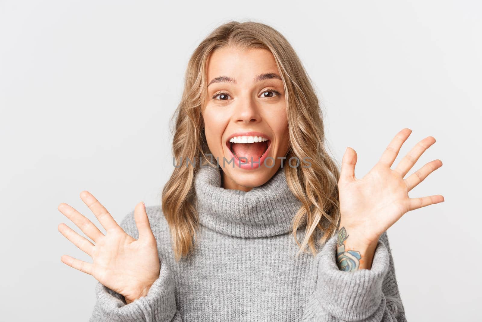 Close-up of friendly attractive girl in grey sweater, smiling and saying hi, waving hand to greet someone, standing over white background by Benzoix