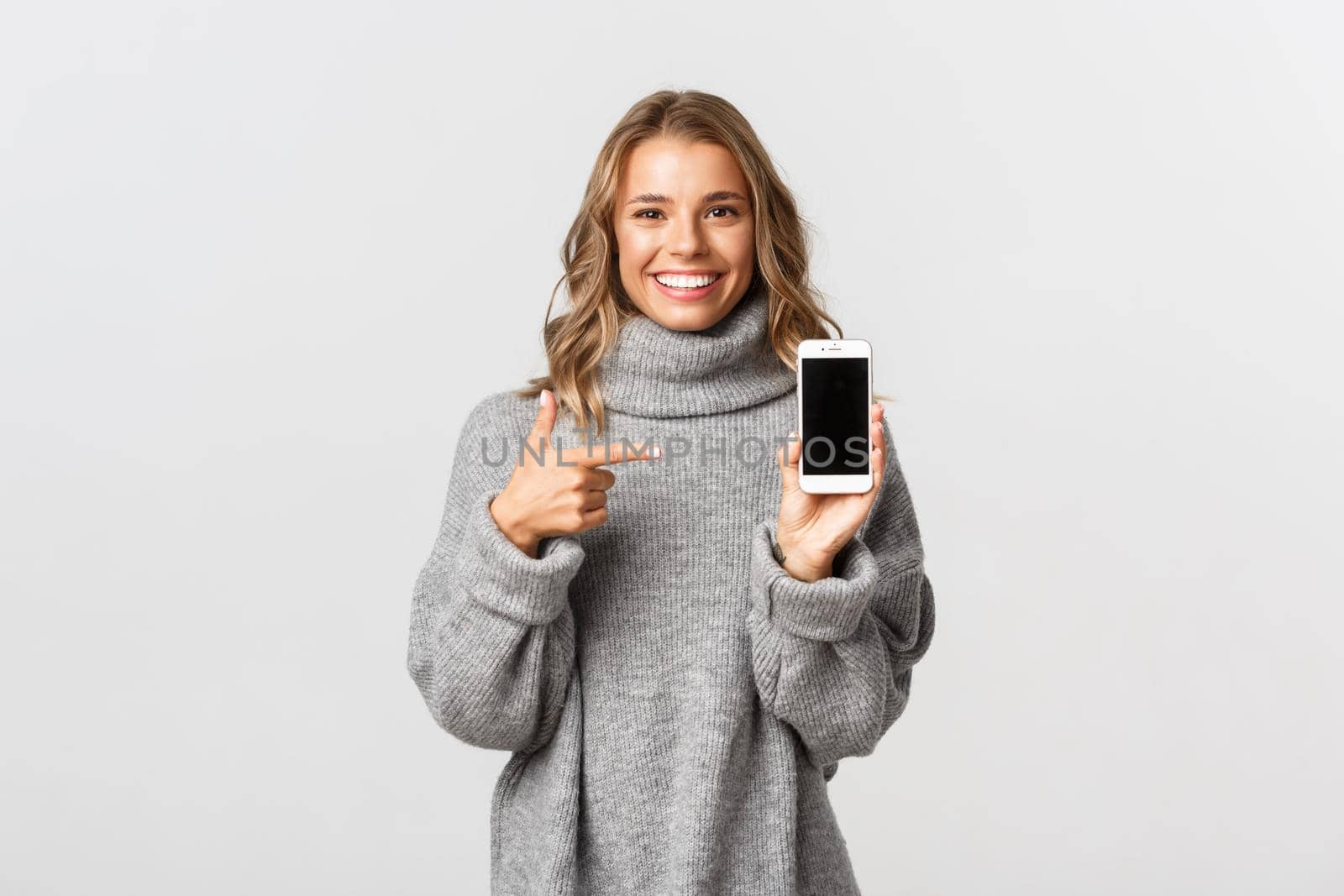 Portrait of attractive blond girl in grey sweater, pointing finger at mobile phone screen and smiling, recommending something by Benzoix