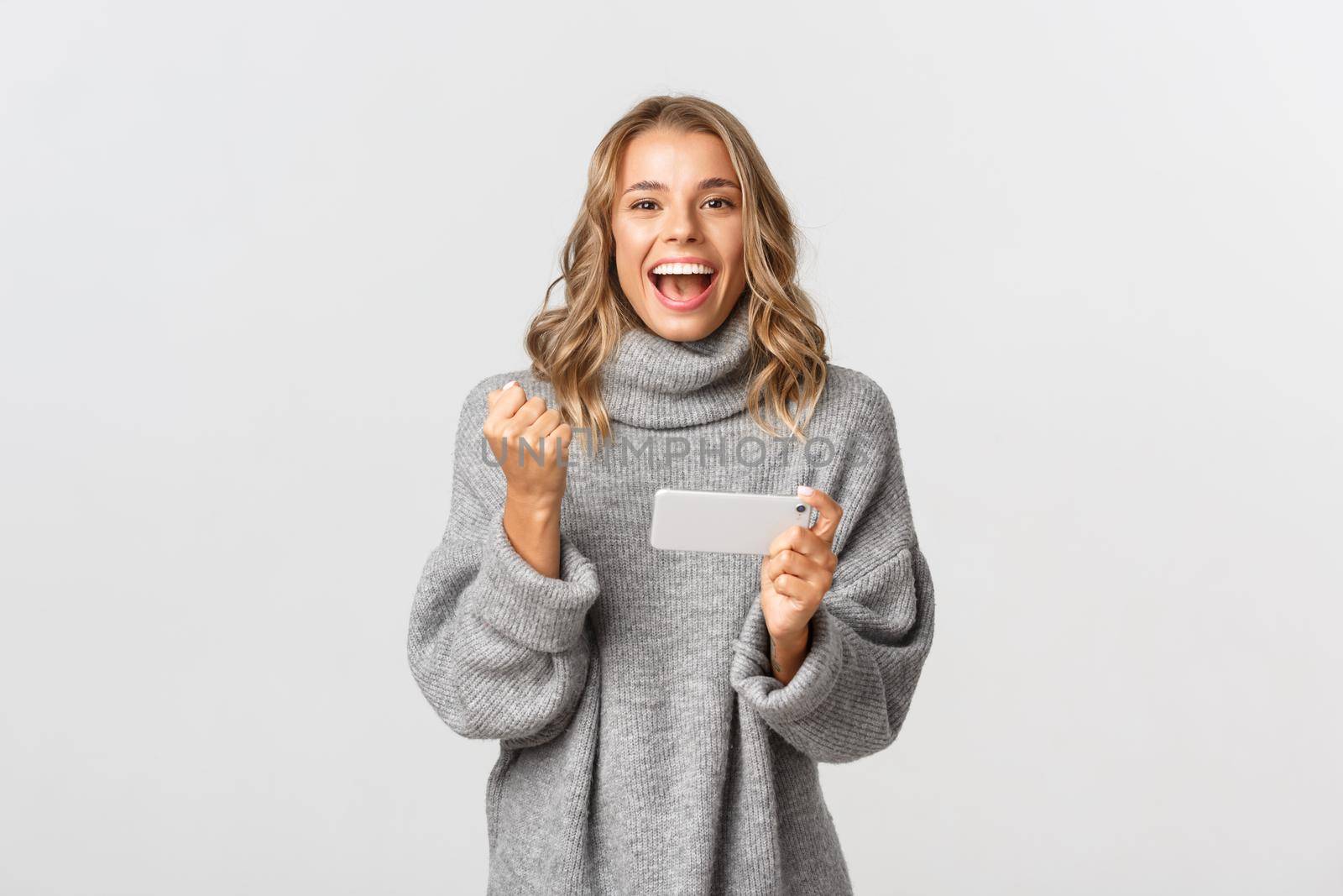 Image of happy winning girl in grey sweater, saying yes and triumphing, holding mobile phone, achieve goal in smartphone application, standing over white background.