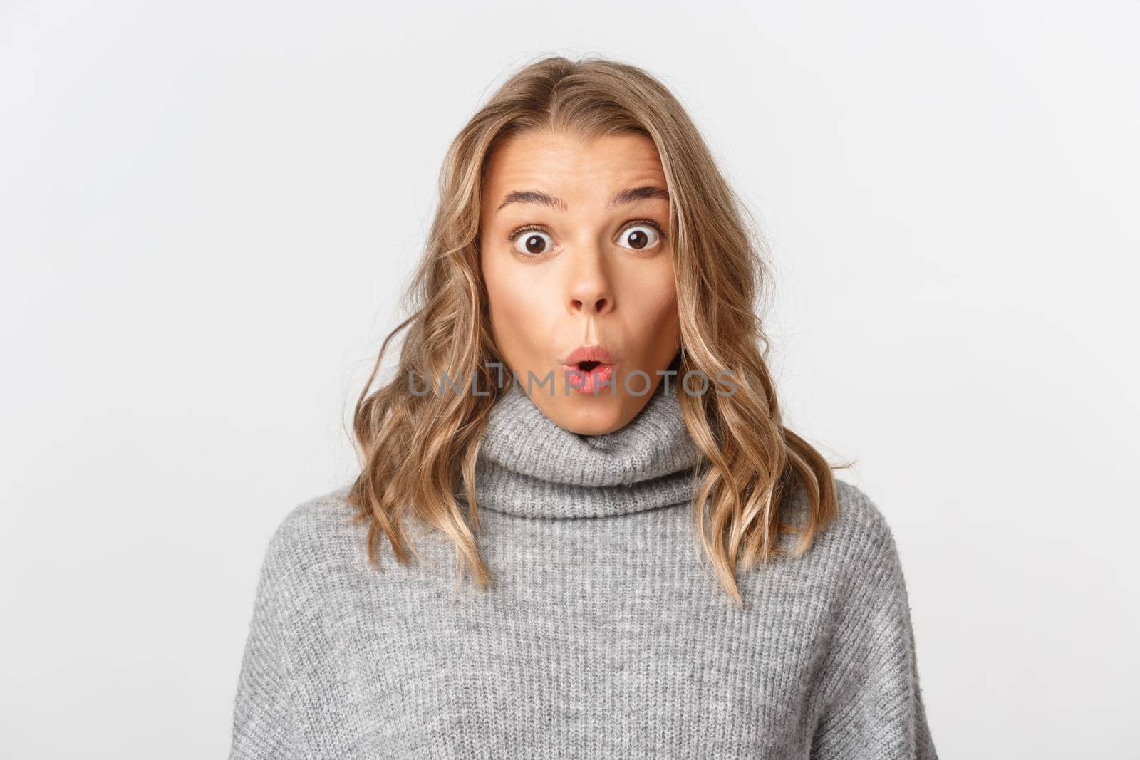 Close-up of attractive blond girl looking surprised, gasping amazed at camera, standing over white background.
