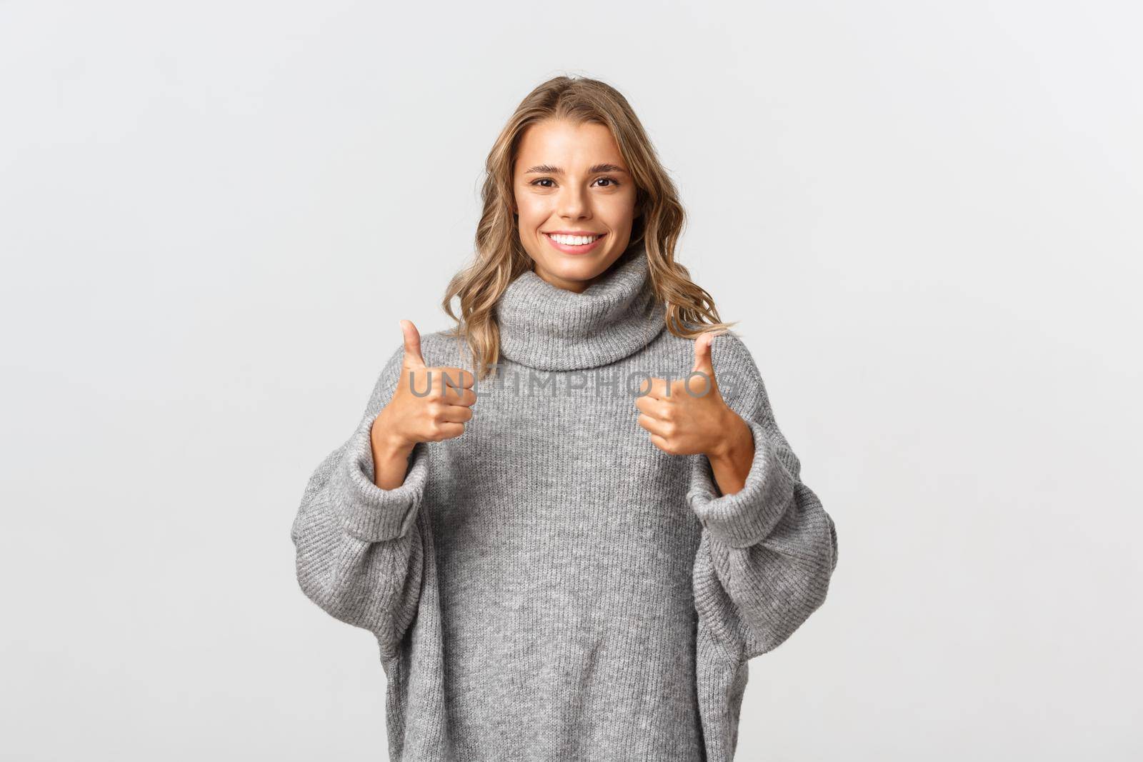 Young supportive woman with blond short hair, showing thumbs-up and smiling, like something or give approval, standing over white background.