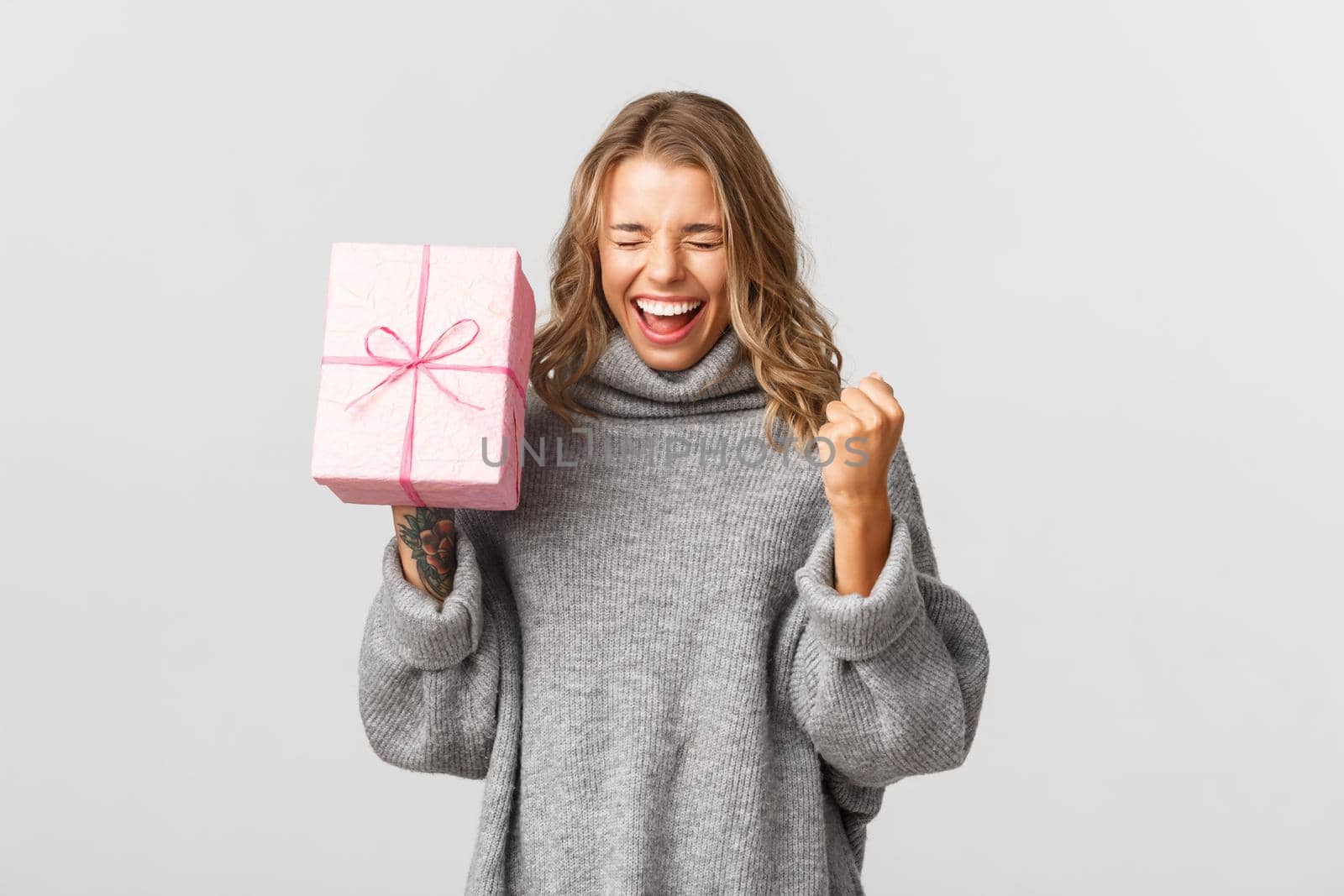 Cheerful pretty girl in grey sweater, looking happy, celebrating her birthday and receiving gift, standing over white background.