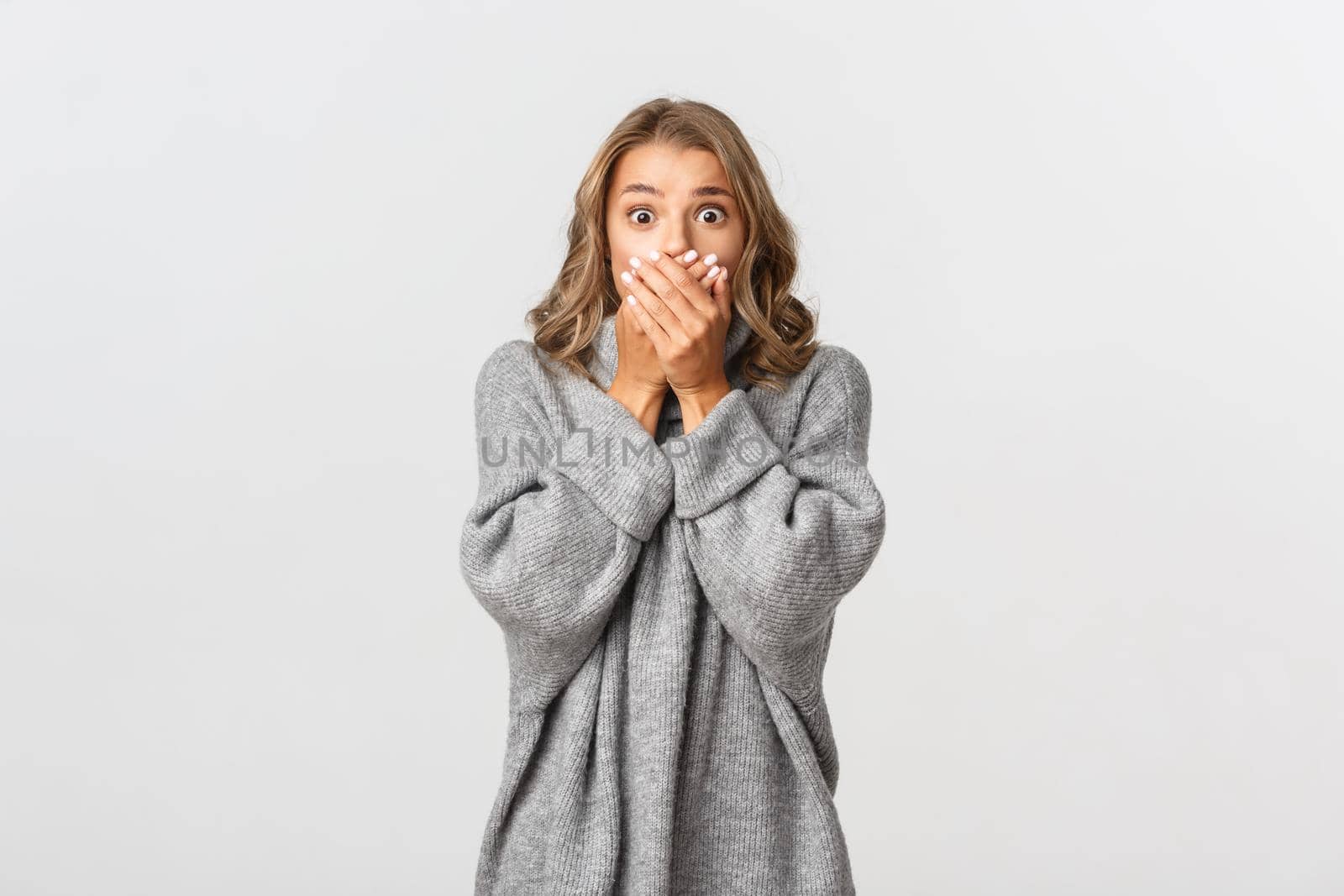 Portrait of blond girl looking shocked and scared at something alarming, gasping and cover mouth, standing over white background.