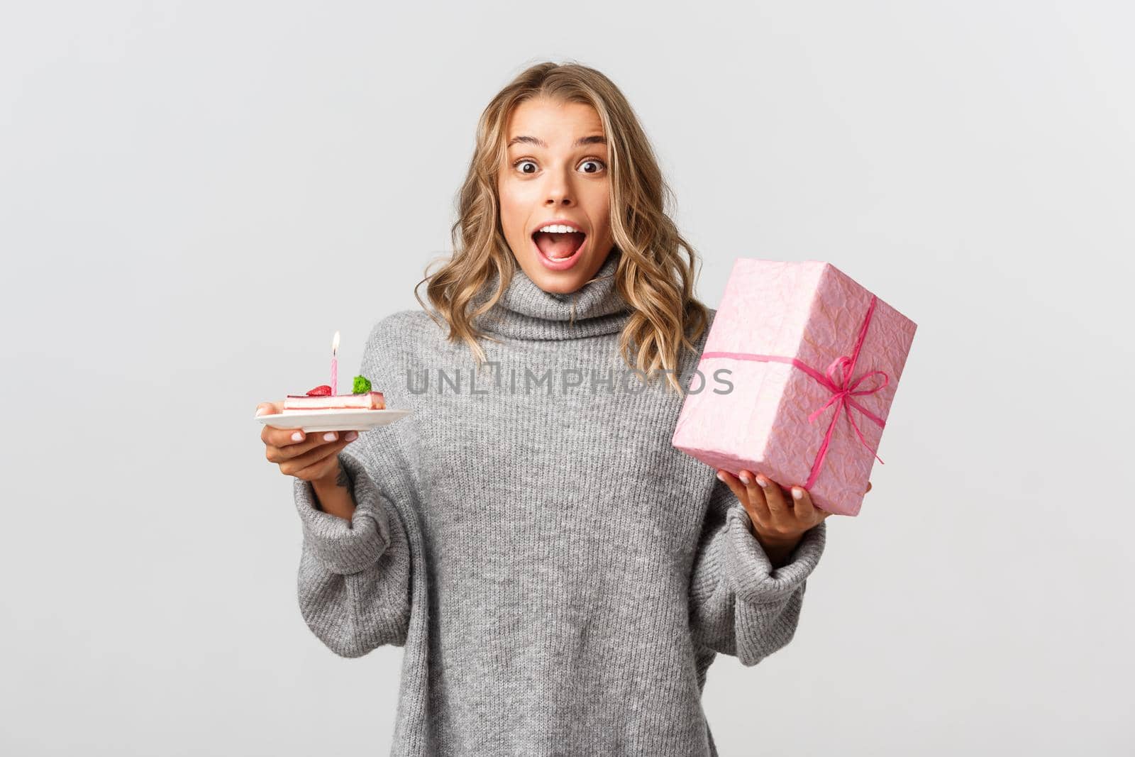 Image of attractive blond girl celebrating birthday, looking surprised, holding b-day cake and a gift, standing over white background.
