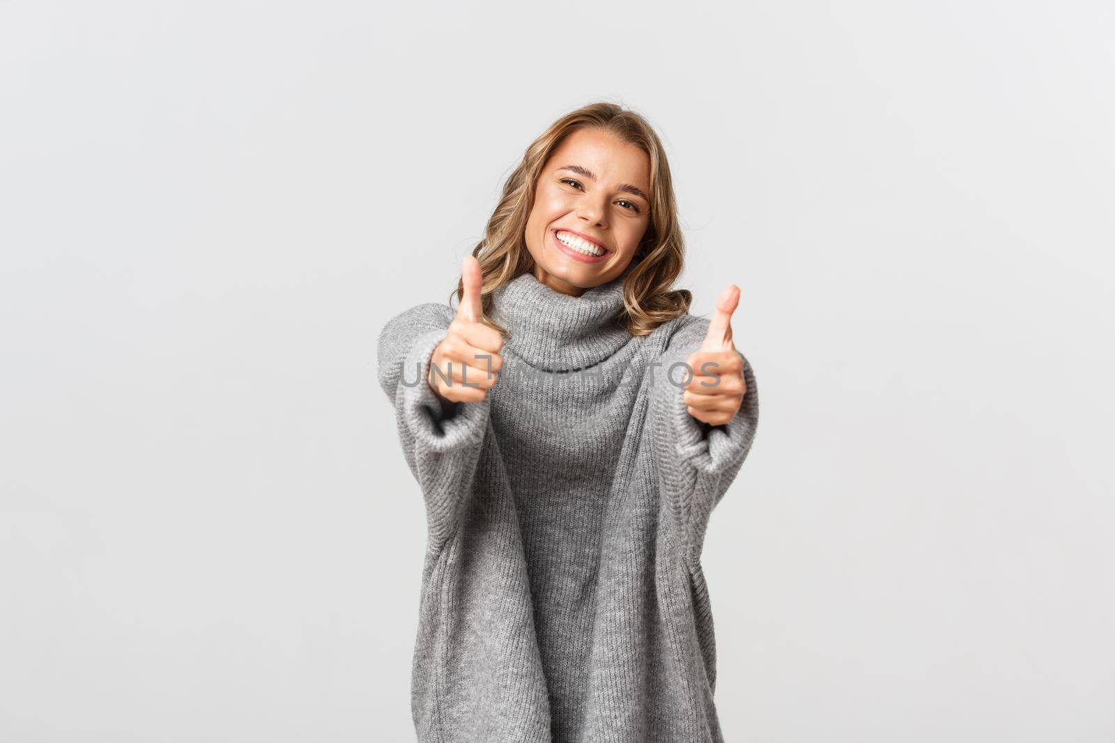 Image of happy beautiful woman in grey sweater, showing thumbs-up and smiling, give approval or agree, like and praise good choice, standing over white background.