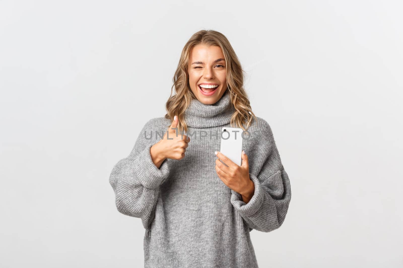 Portrait of attractive blond girl in grey sweater winking satisfied, showing thumbs-up and using mobile phone, recommend smartphone app, standing over white background.