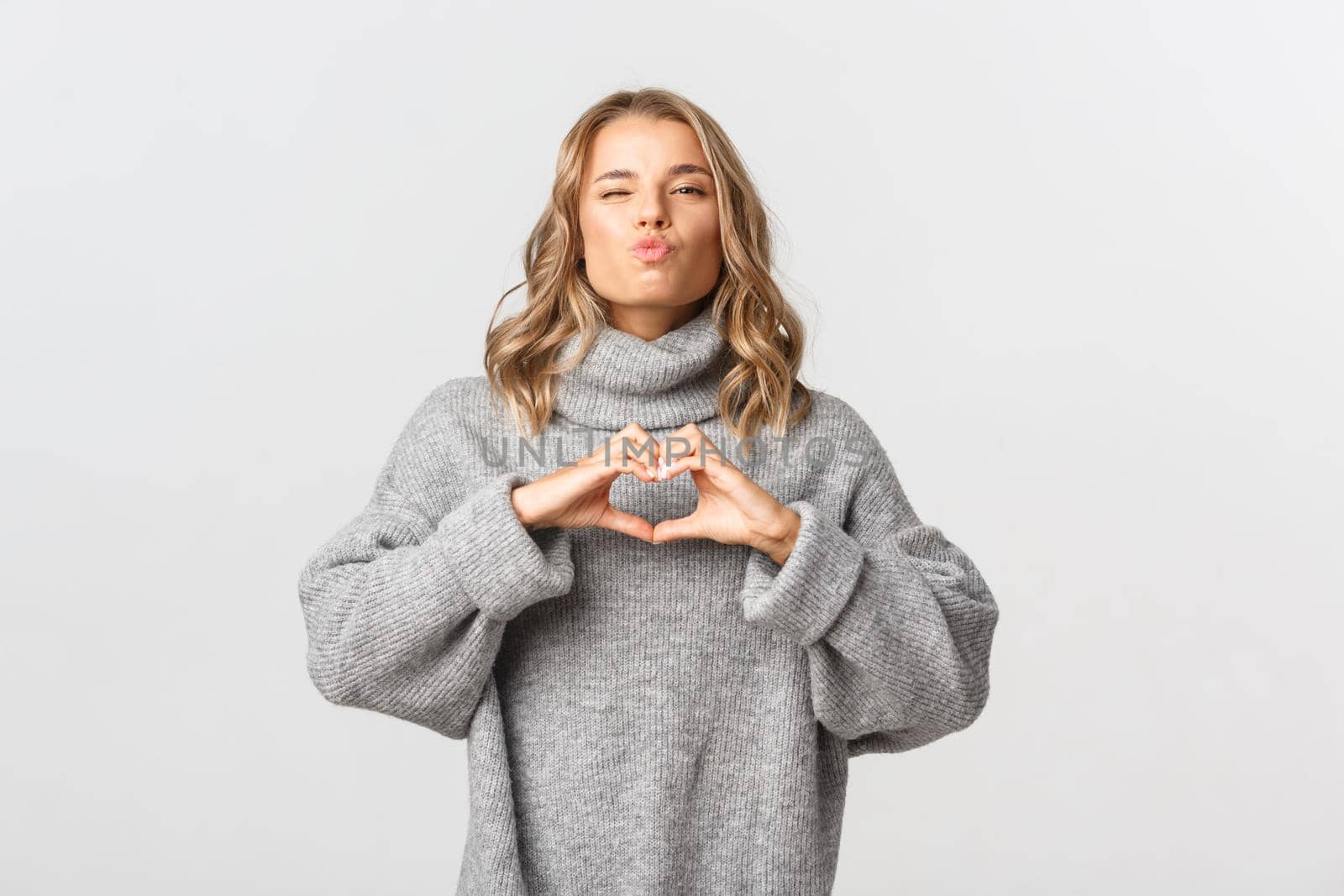Image of attractive tender girl in grey sweater, confess in love, showing heart sign and waiting for kiss with eyes closed, standing over white background.