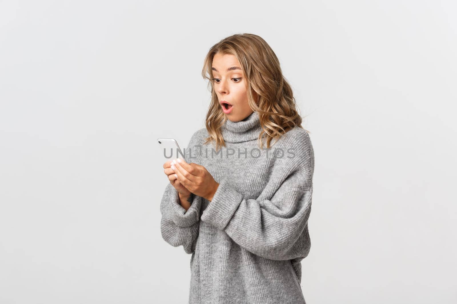 Portrait of amazed blond girl looking excited at smartphone screen, standing over white background. Concept of technology and communication.