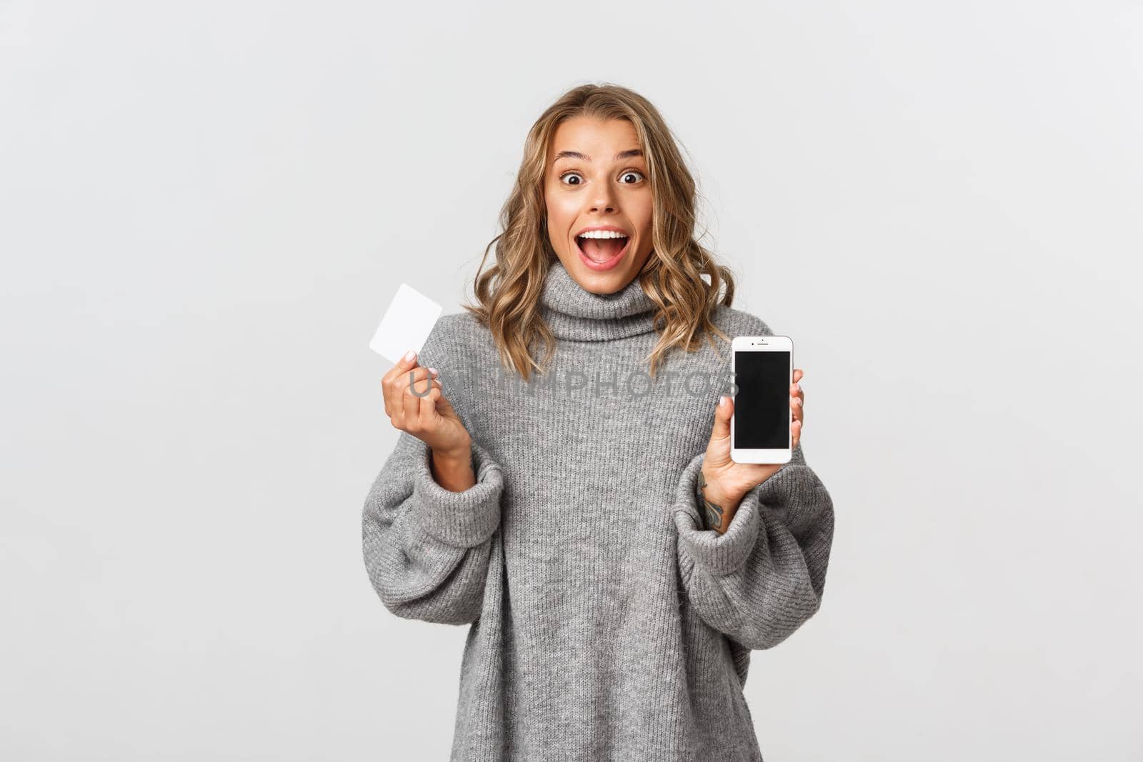 Excited beautiful girl with blond short hairstyle, showing smartphone screen and holding credit card, standing over white background by Benzoix