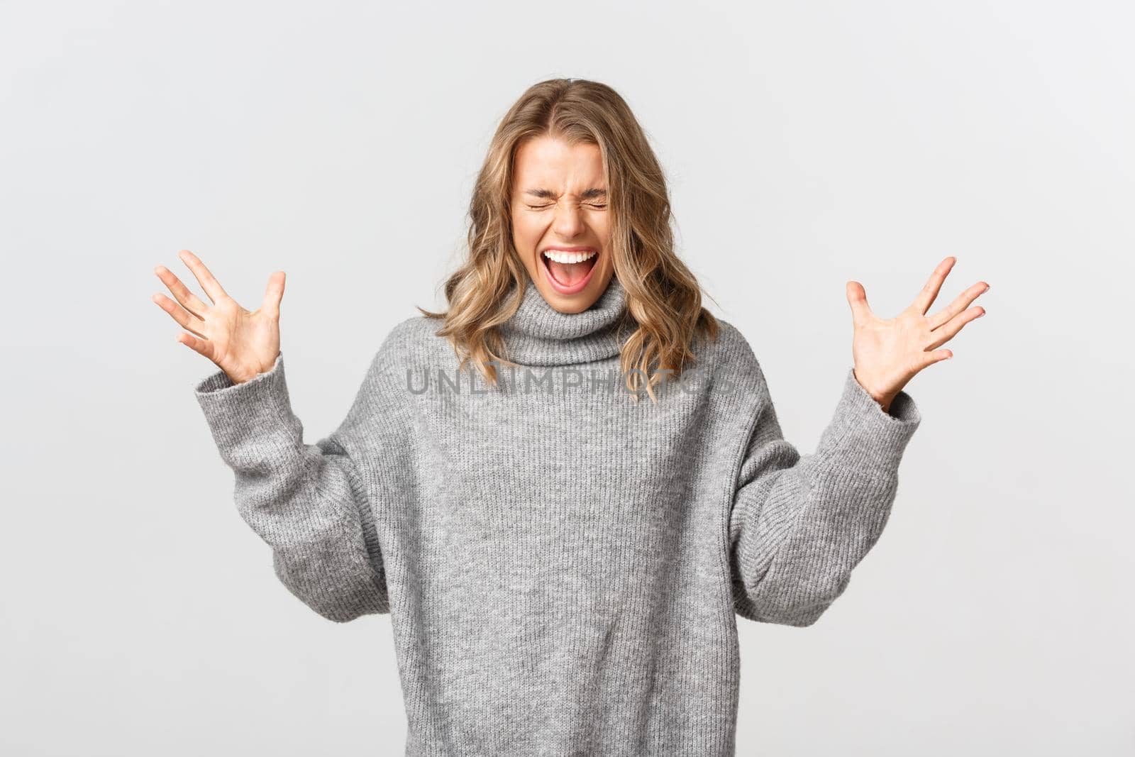 Portrait of blond girl in casual grey sweater losing control over emotions, screaming and shaking hands distressed, standing over white background and yelling.