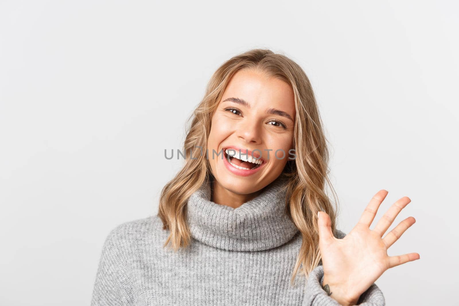 Close-up of friendly attractive girl in grey sweater, smiling and saying hi, waving hand to greet someone, standing over white background by Benzoix