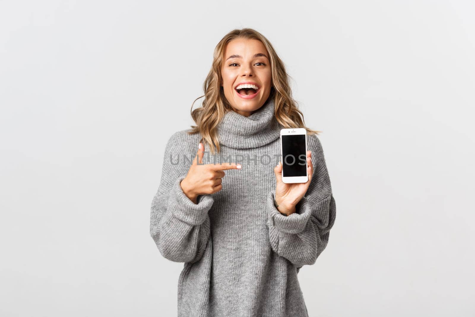Portrait of happy attractive caucasian girl in grey sweater, laughing at something funny, pointing finger at smartphone screen, standing over white background.