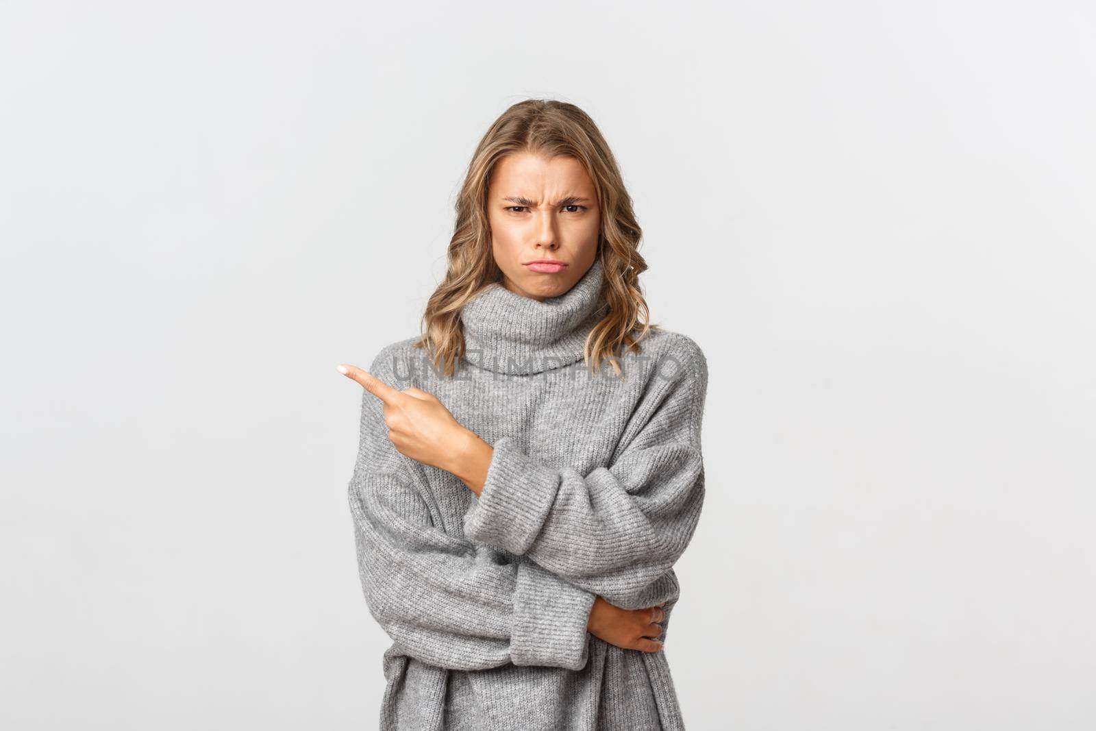 Image of disappointed moody girl, wearing grey sweater, sulking and frowning upset, pointing finger at upper left corner, standing over white background by Benzoix
