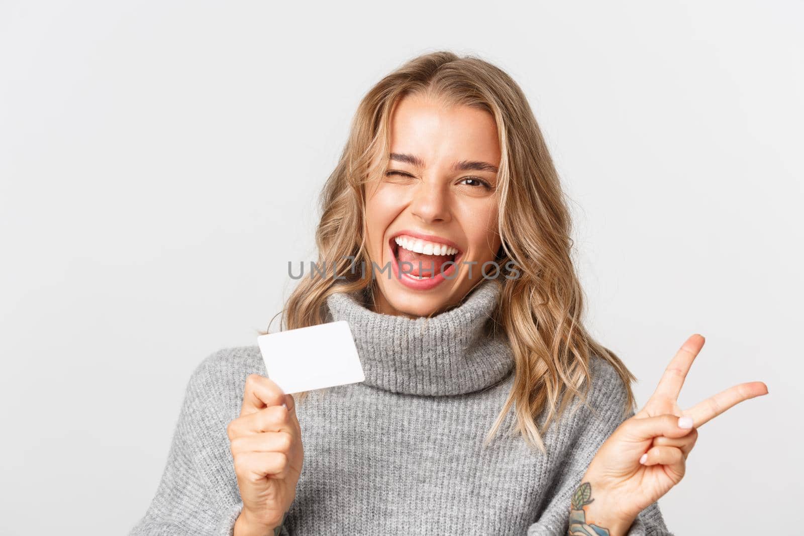 Close-up of attractive blond girl in grey sweater, showing credit card and peace sign, standing over white background happy.