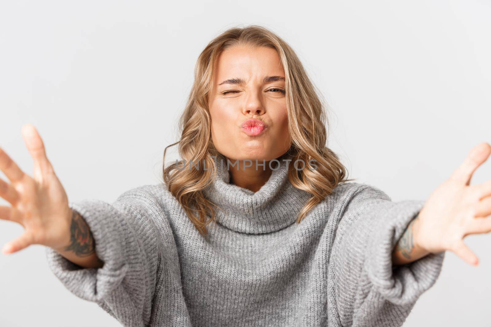 Close-up of happy attractive blond girl smiling, stretching hands forward for hug, reaching for something with kissing face, standing over white background.