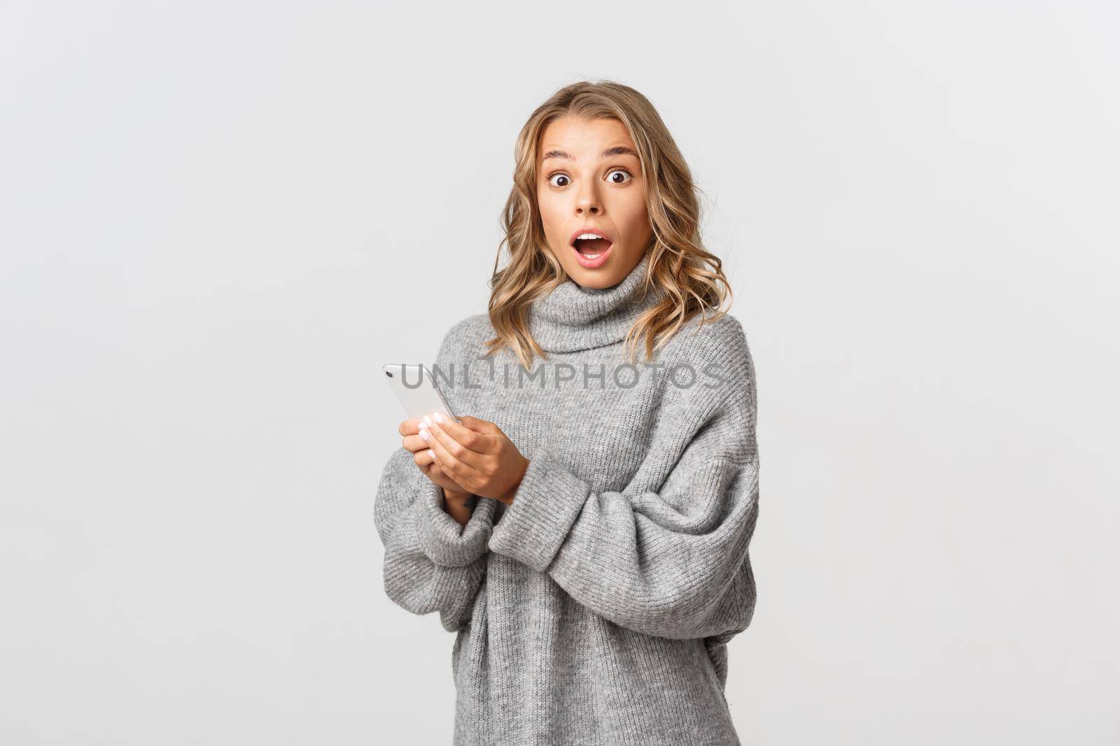 Portrait of attractive blond girl gasping surprised, holding smartphone. Concept of online shopping and technology.
