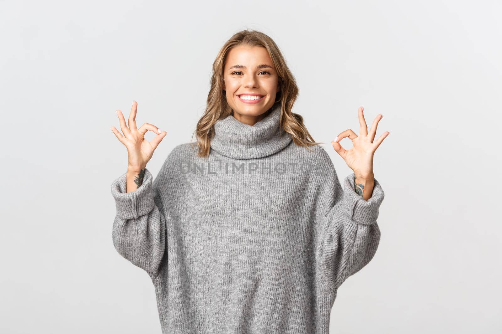 Young satisfied woman with blond short hair, wearing grey sweater, showing okay signs and smiling pleased, standing over white background.