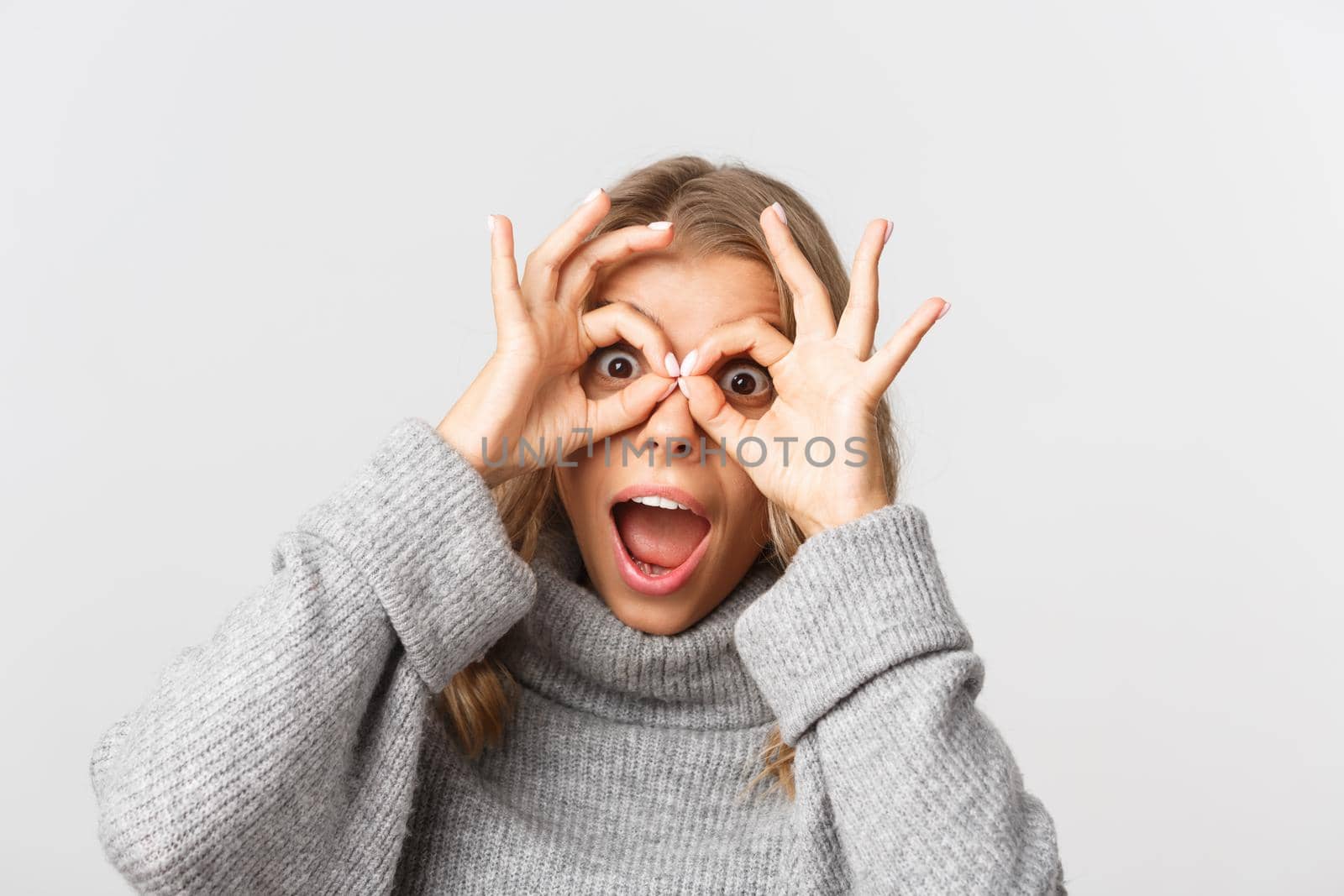 Close-up of happy attractive girl having fun, looking through finger glasses and look amazed, standing over white background.