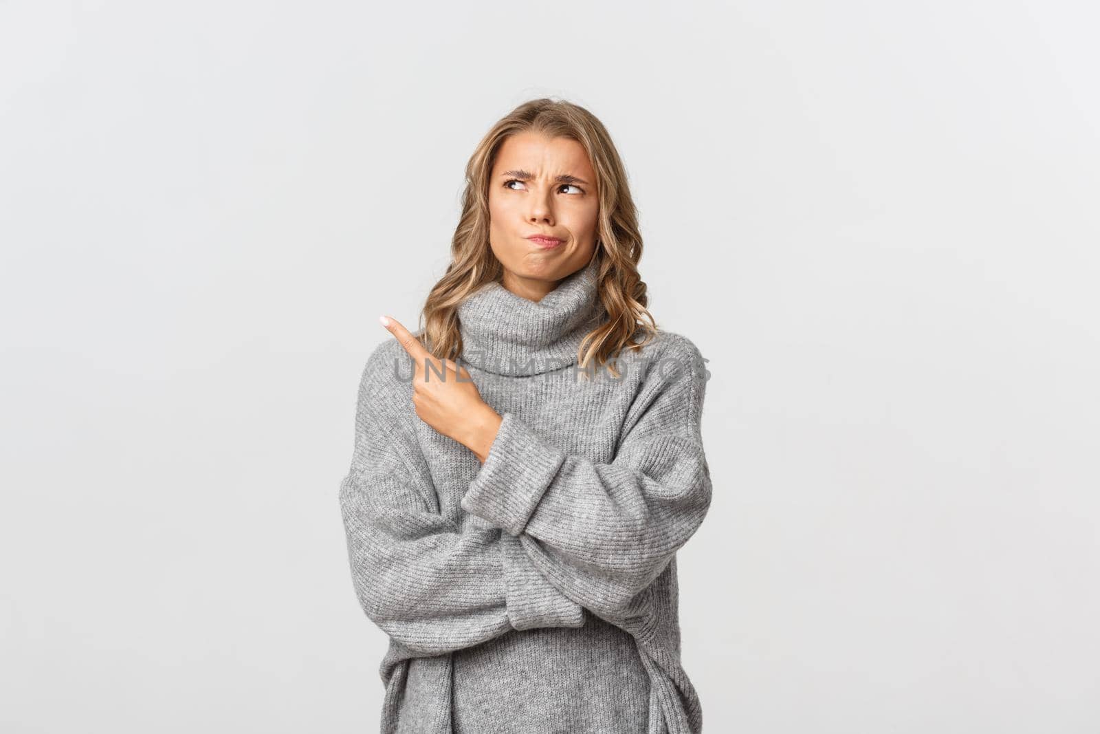 Image of skeptical blond girl in grey sweater, looking indecisive and pointing at upper left corner, smirking doubtful, standing over white background.
