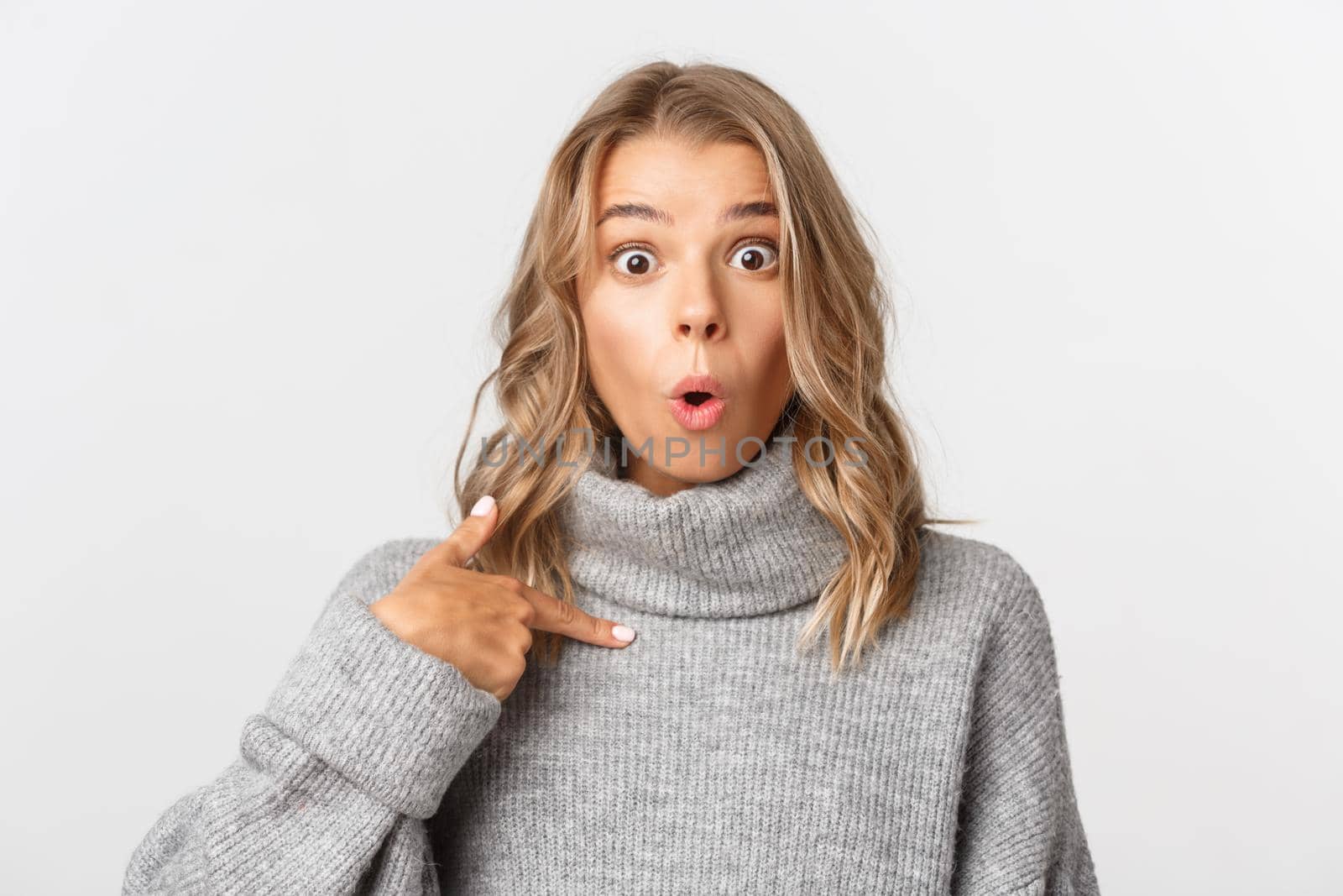 Close-up of attractive surprised girl pointing at herself, looking wondered, standing over white background.