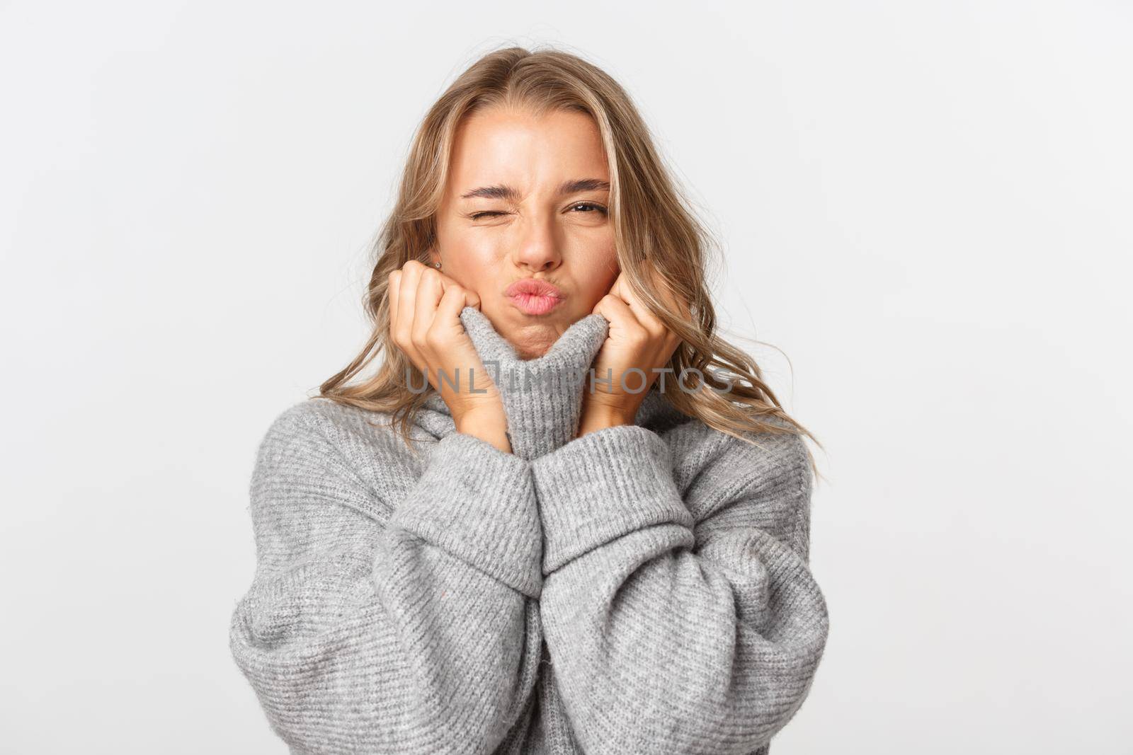 Close-up of sassy blond girl in grey warm sweater, pouting for kiss and squinting coquettish, standing over white background.