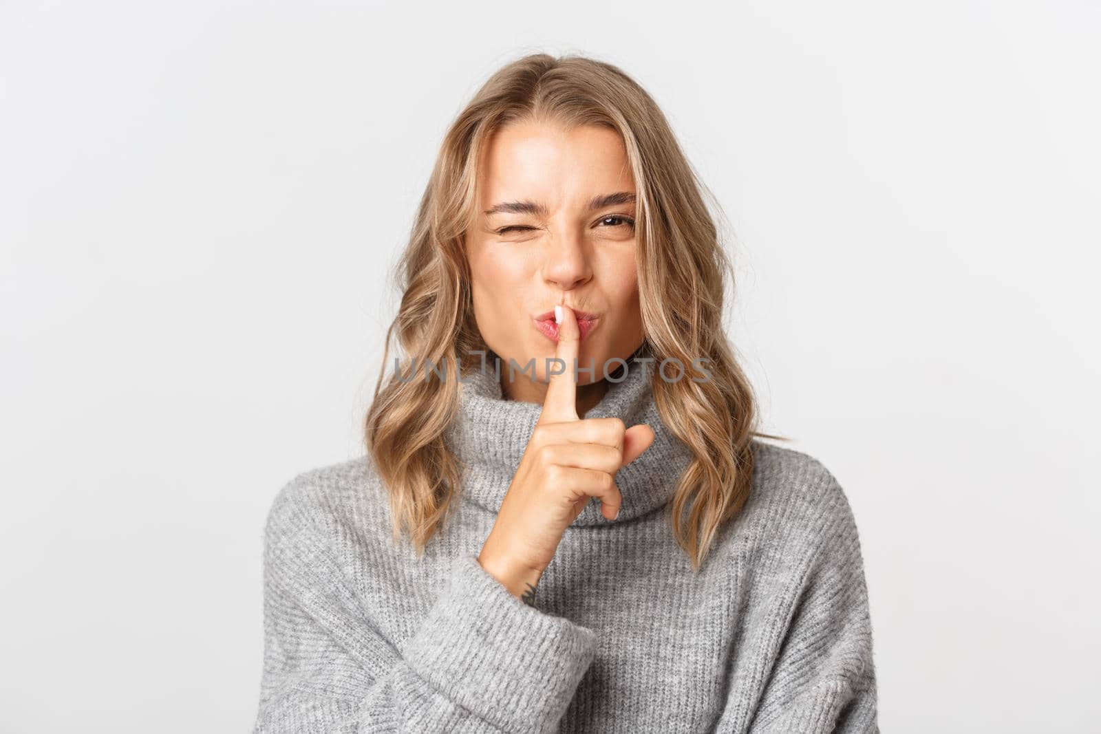 Close-up of attractive blond girl in grey sweater sharing a secret, shushing at winking, asking to be quiet, standing over white background by Benzoix