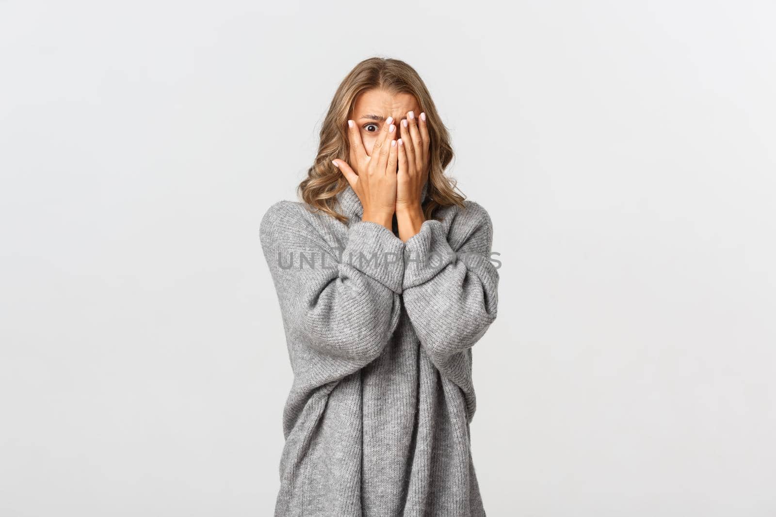 Image of scared woman in grey sweater, shut her eyes but peeking through fingers at something scary, standing horrified over white background.