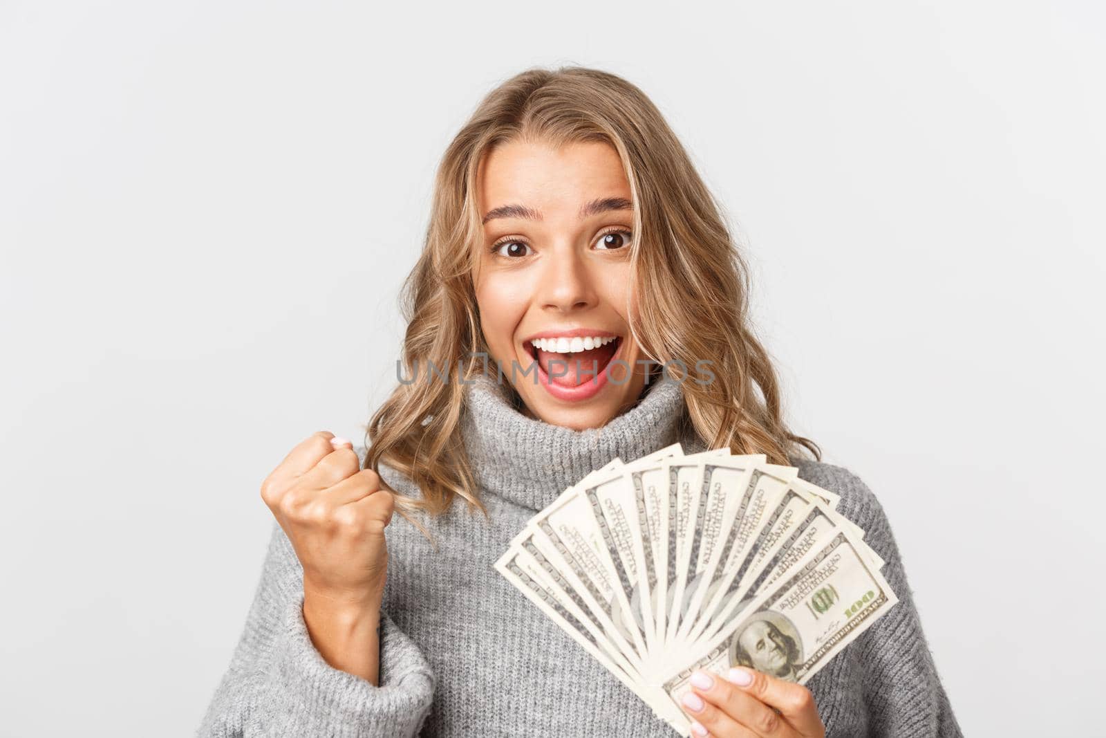 Close-up of successful and rich blond girl in grey sweater, holding money and looking like winner, standing over white background.