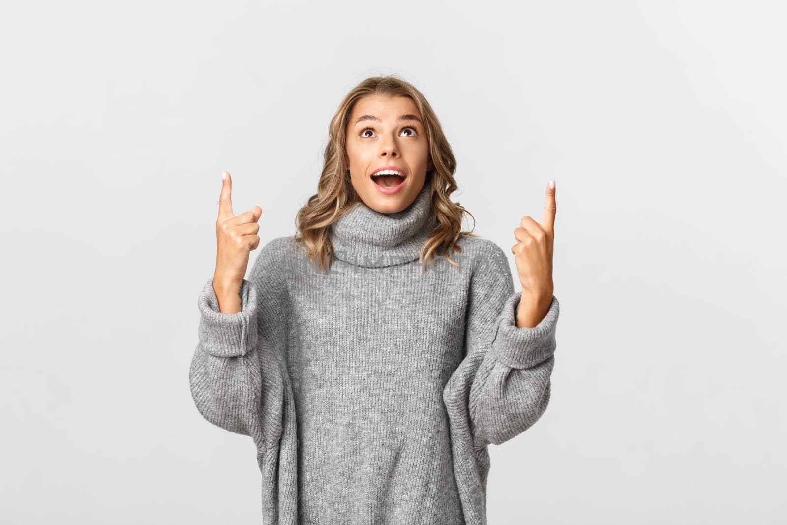 Image of surprised and happy blond girl, wearing grey sweater, pointing fingers up and looking fascinated, white background.