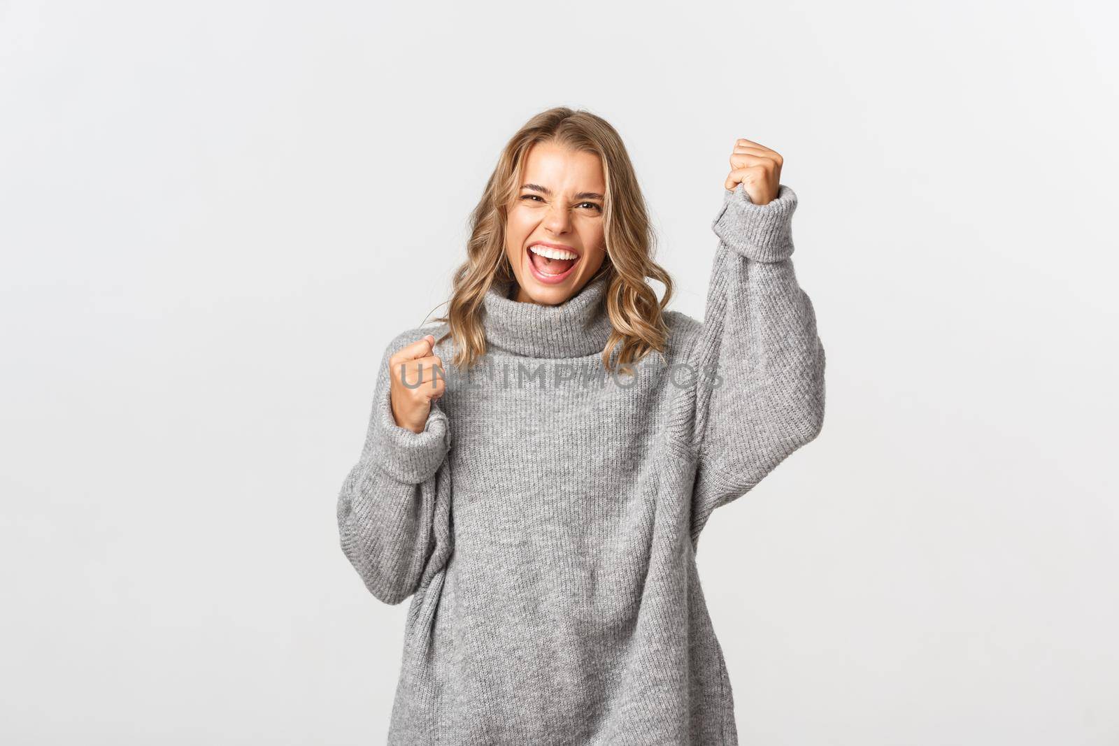 Close-up of excited pretty girl in grey sweater, cheering and rooting for something, screaming for joy, raising hand up and triumphing, standing over white background.