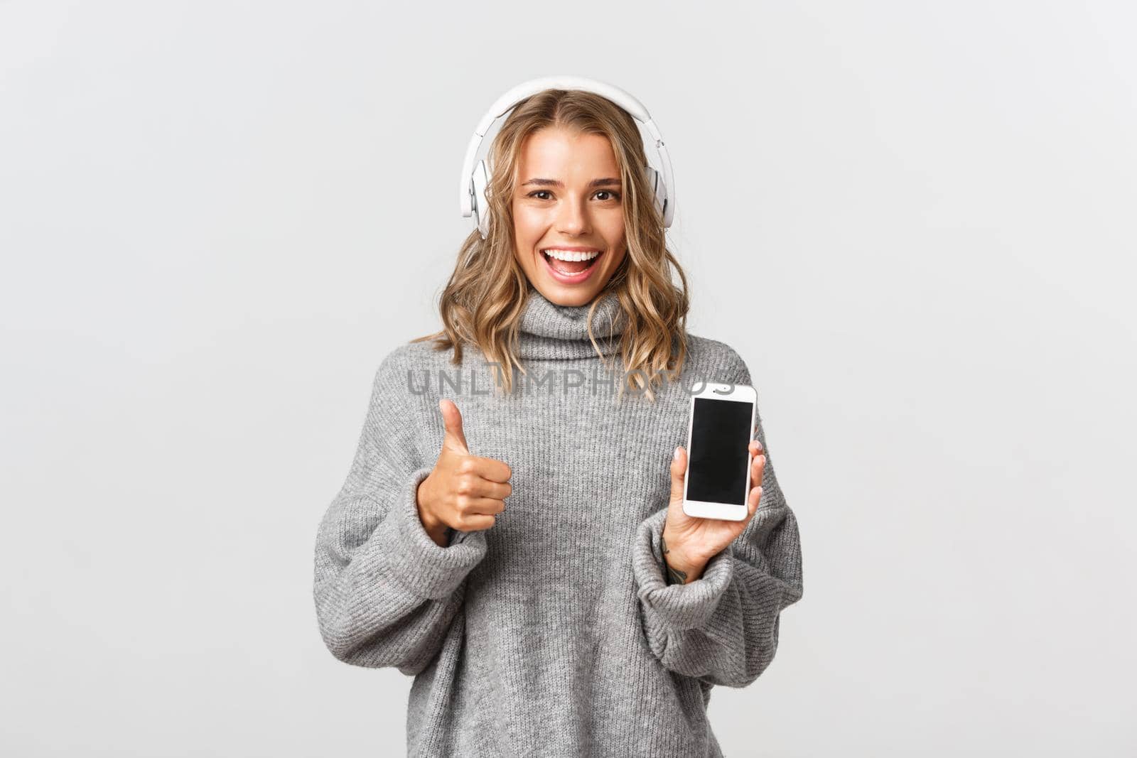 Image of satisfied blond girl looking happy, showing thumbs-up, listening music in headphones and demonstrate smartphone screen, white background.