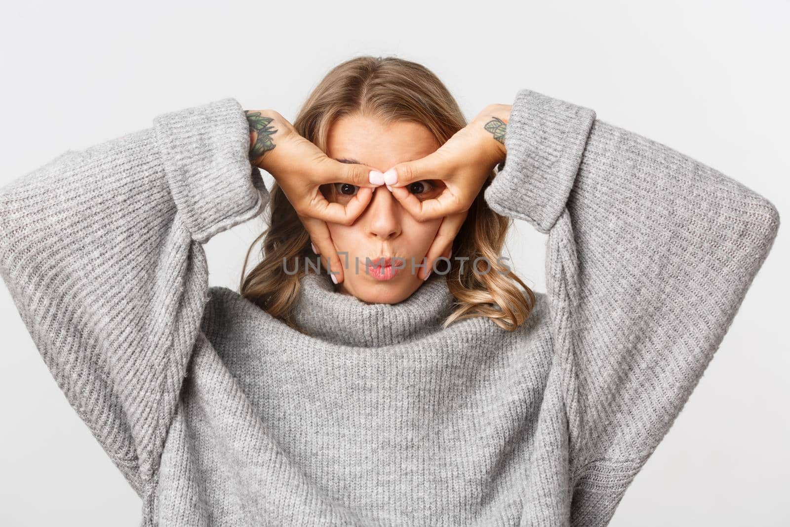 Close-up of funny blond girl in grey sweater, showing superhero mask with hands and pouting silly, standing over white background by Benzoix
