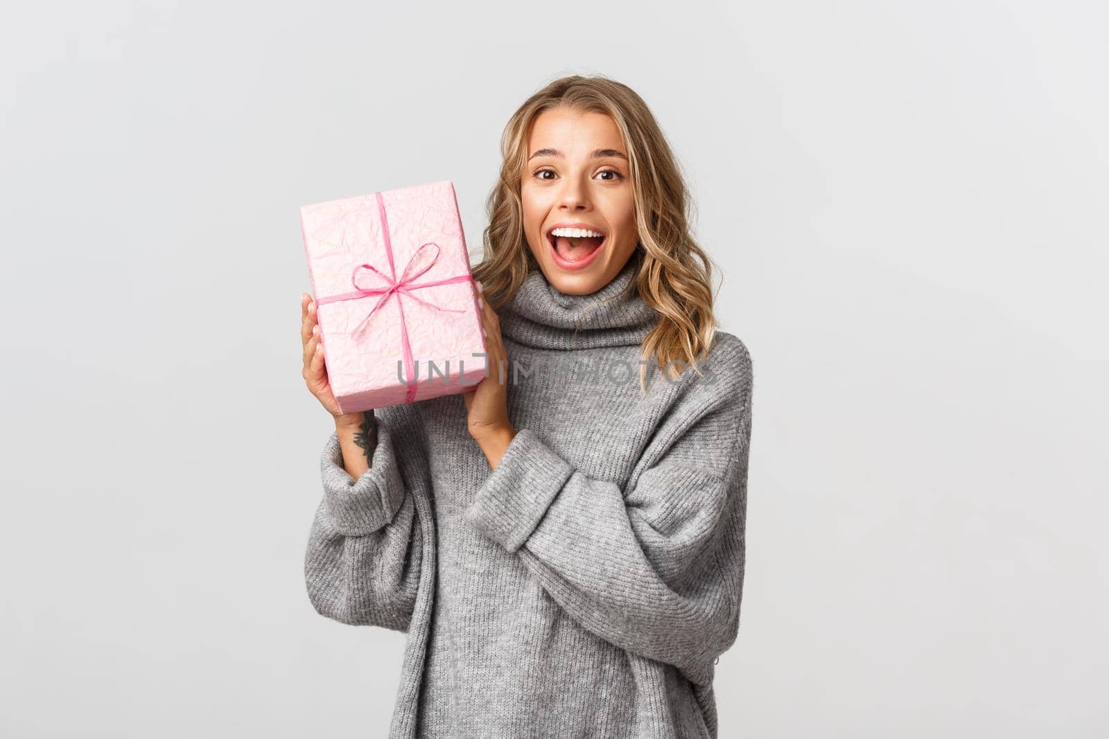 Portrait of attractive blond girl in grey sweater, looking happy, receiving birthday gift in pink box, standing over white background.