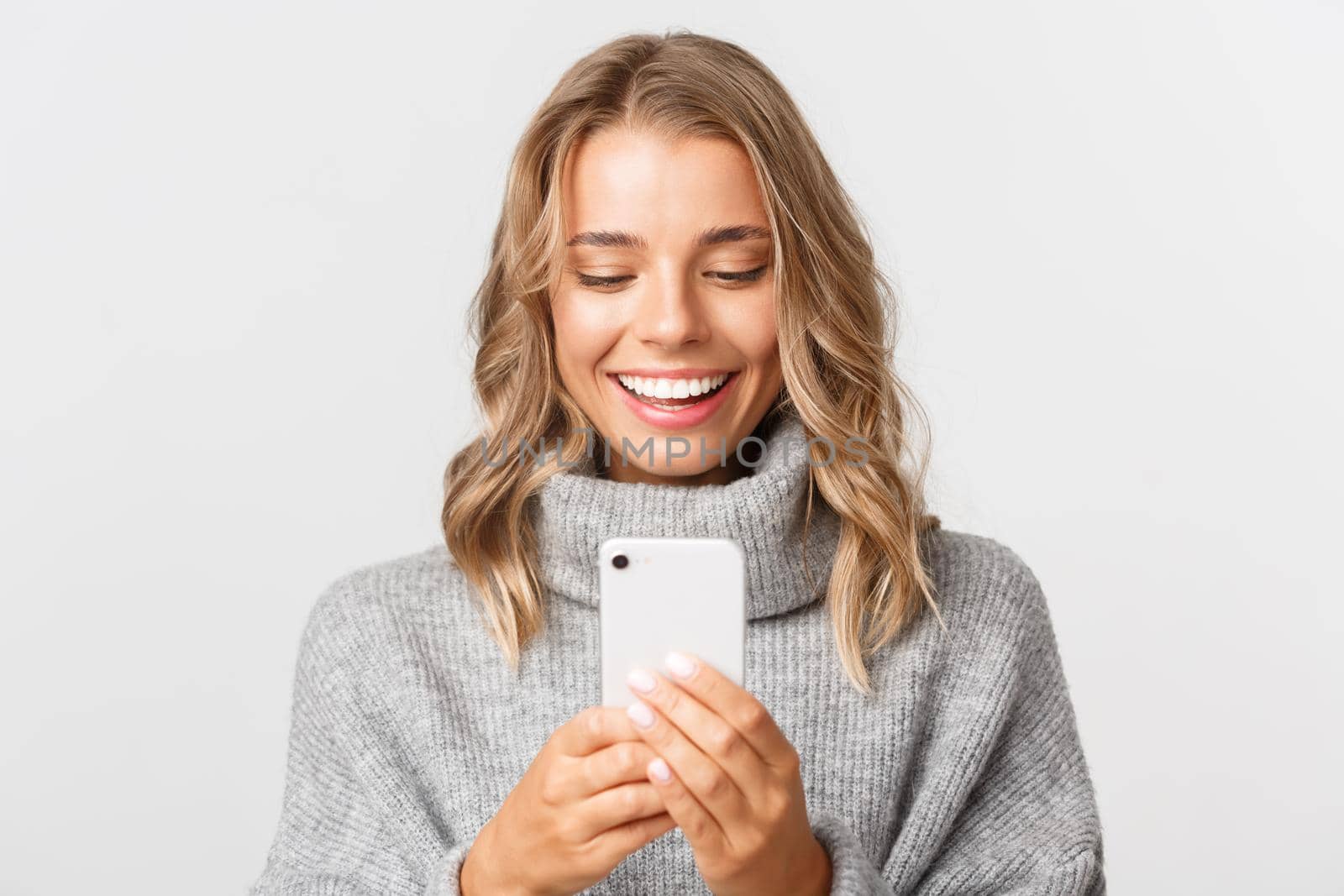 Close-up of attractive blond girl in grey sweater, checking messages on phone or shopping online, looking at smartphone with happy face, standing over white background.