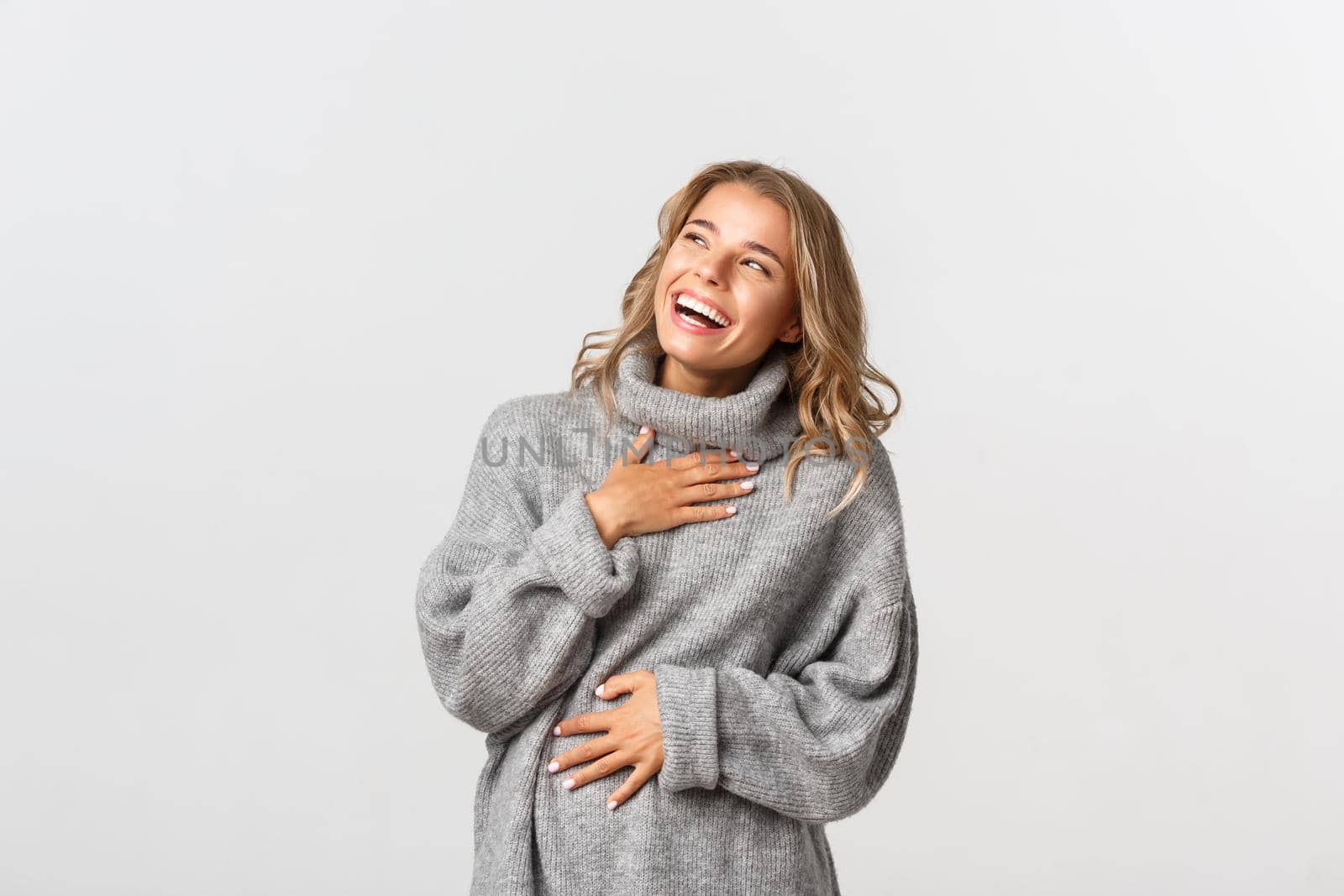 Close-up of happy beautiful woman with short blond hairstyle, laughing and looking at upper left corner, standing in grey sweater, white background.