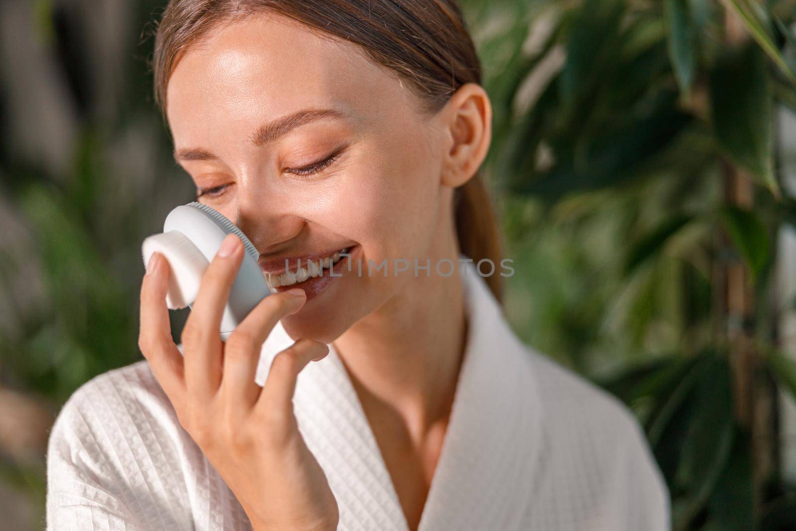 Portrait of young woman in bathrobe smiling and holding silicone face brush for skin exfoliating, enjoying beauty routine by Yaroslav_astakhov