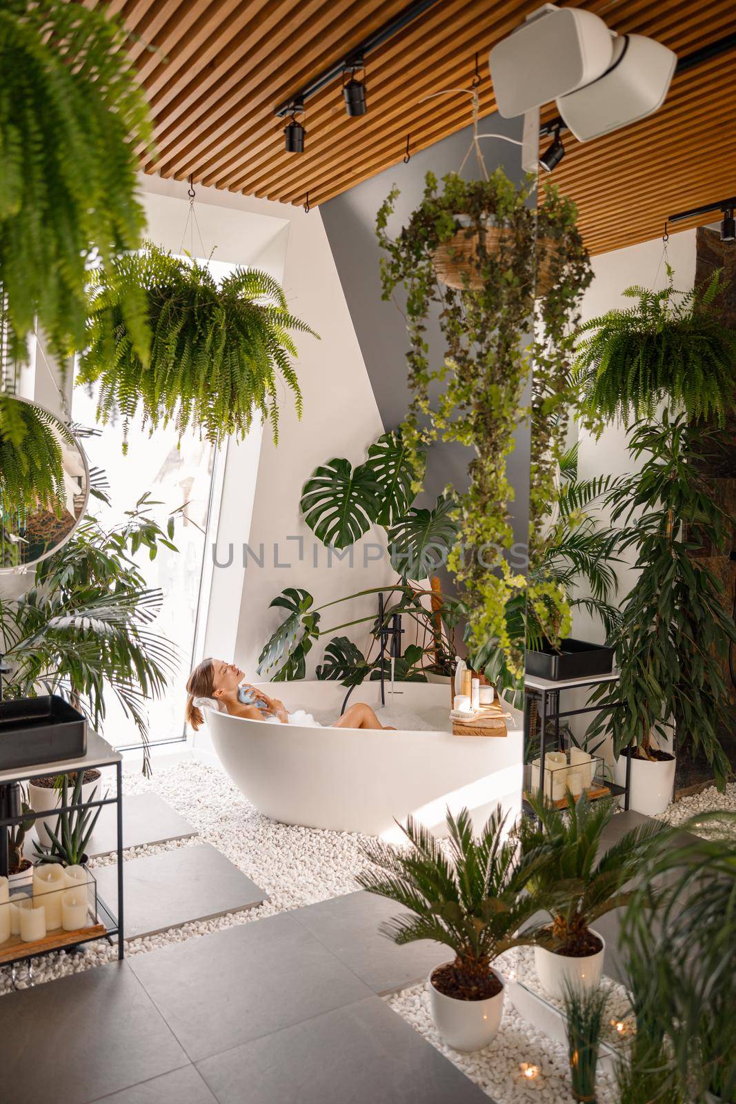 Relaxed young woman taking bubble bath in modern bathroom decorated with tropical plants at luxury spa resort. Wellness, body care concept