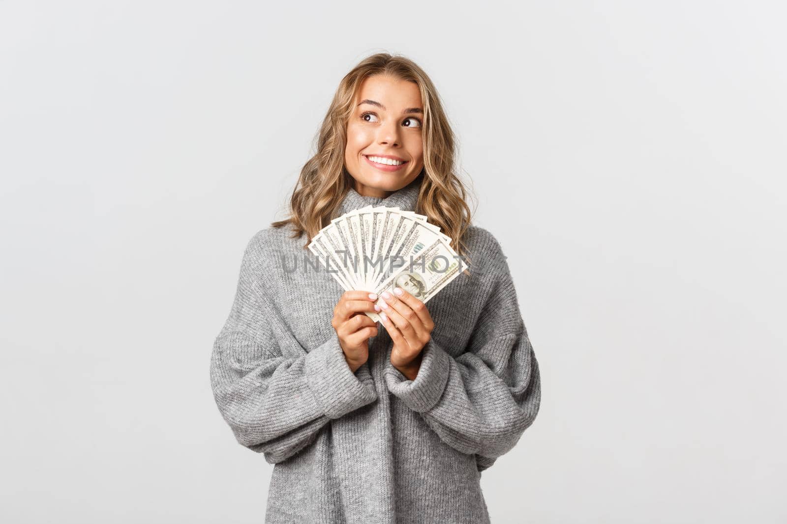 Portrait of dreamy cute girl in grey sweater, looking at upper left corner thoughtful and smiling, holding money, white background.