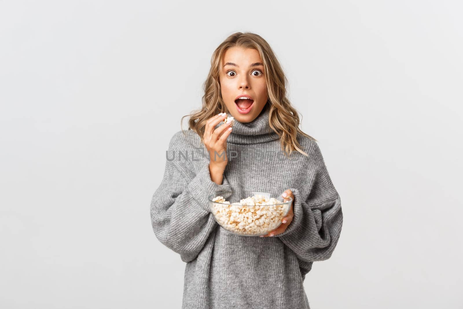 Image of amazed blond girl watching movie, looking excited, eating popcorn, standing over white background.