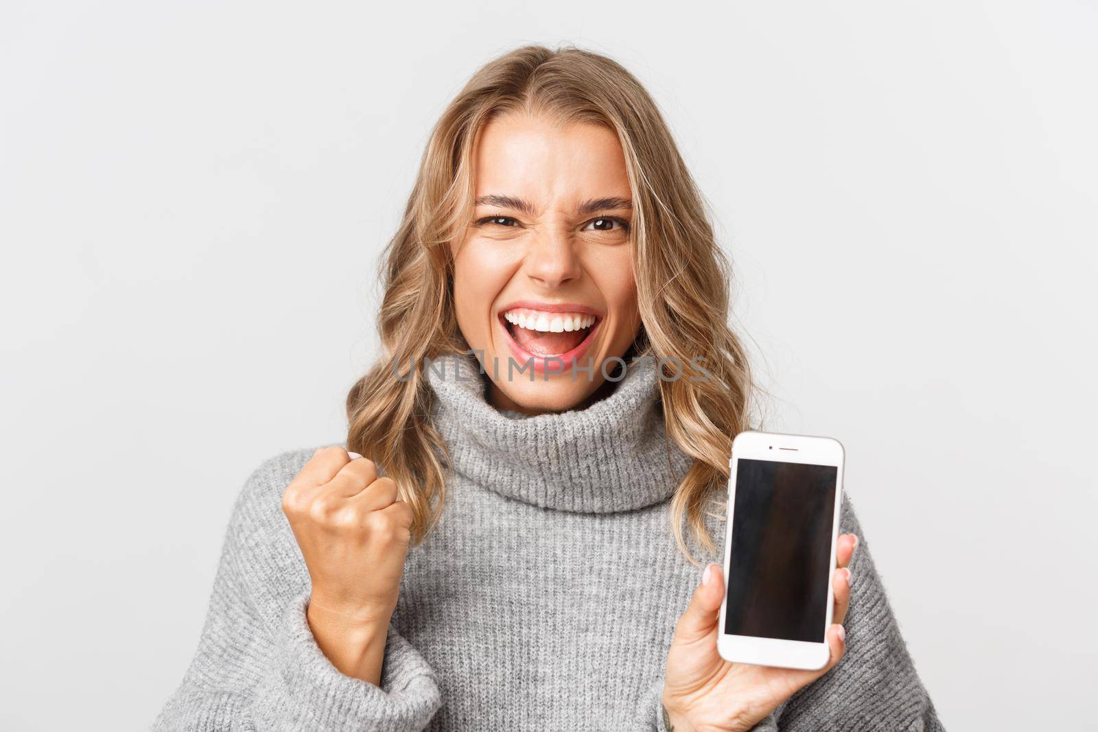 Close-up of successful blond girl, looking happy and triumphing, showing smartphone screen, standing over white background.