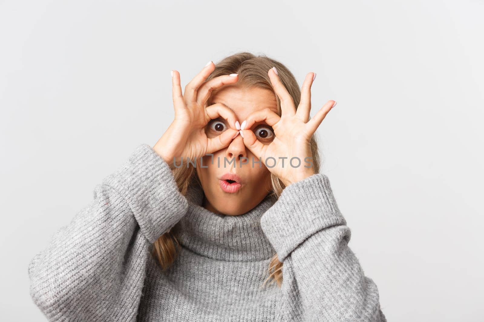 Close-up of beautiful cute girl with blond short hairstyle, making finger glasses and fooling around, standing over white background.